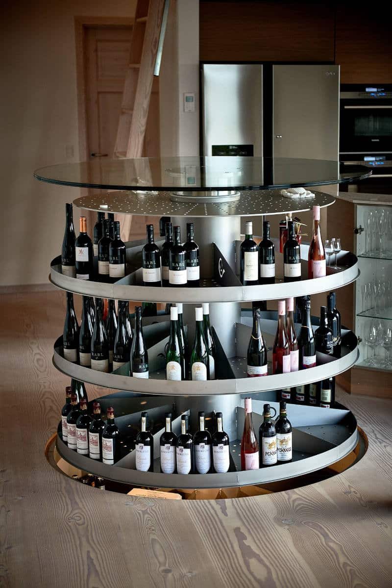 Kælderen hævet i et køkken: runde metalhylder med flasker, glaslåget løftet over.