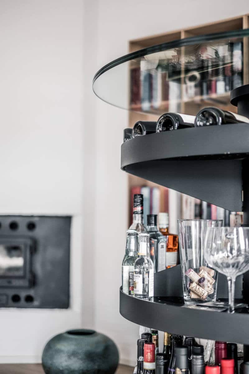 Mat sort metalhylde med flasker, glas og propper. Pejs i baggrunden.